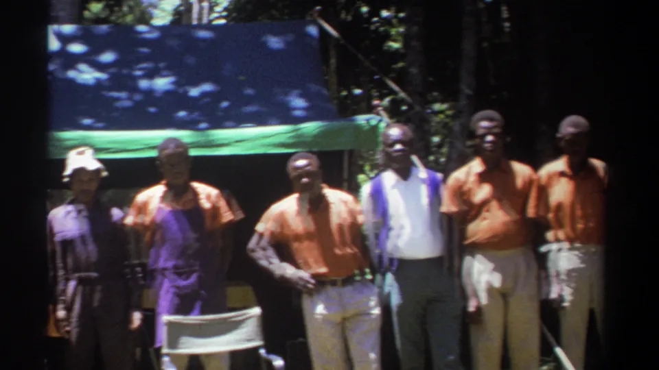 A group of Black men stands near white folding chairs beneath a green tarp in a wooded Kenyan outdoor setting.