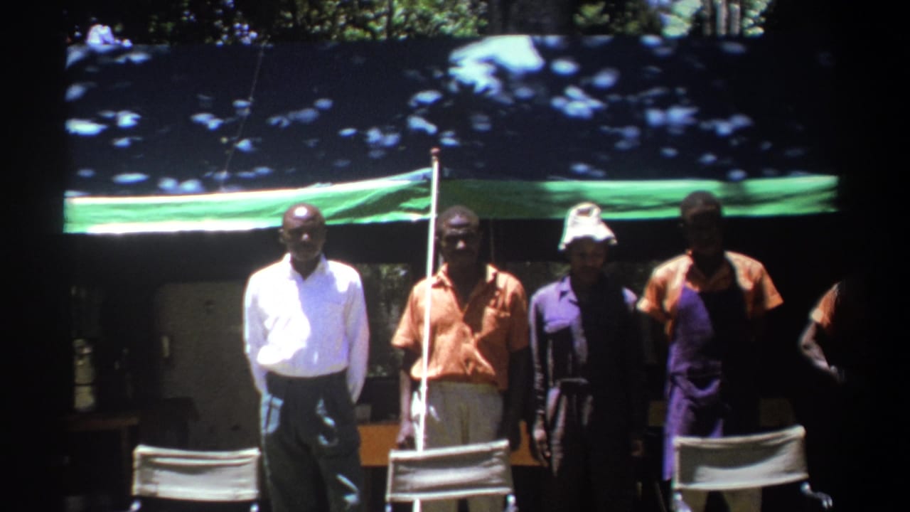 Men stand facing the camera under a tarp, with one man in a white shirt at left.