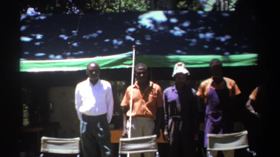 Men face the camera beneath a blue and green tarp in 1969 Kenya, including one in a white shirt and others in work clothes.
