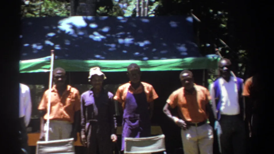 A line of adult men stands near folding chairs under a tarp in Kenya, 1969, with strong sun patches across the scene.