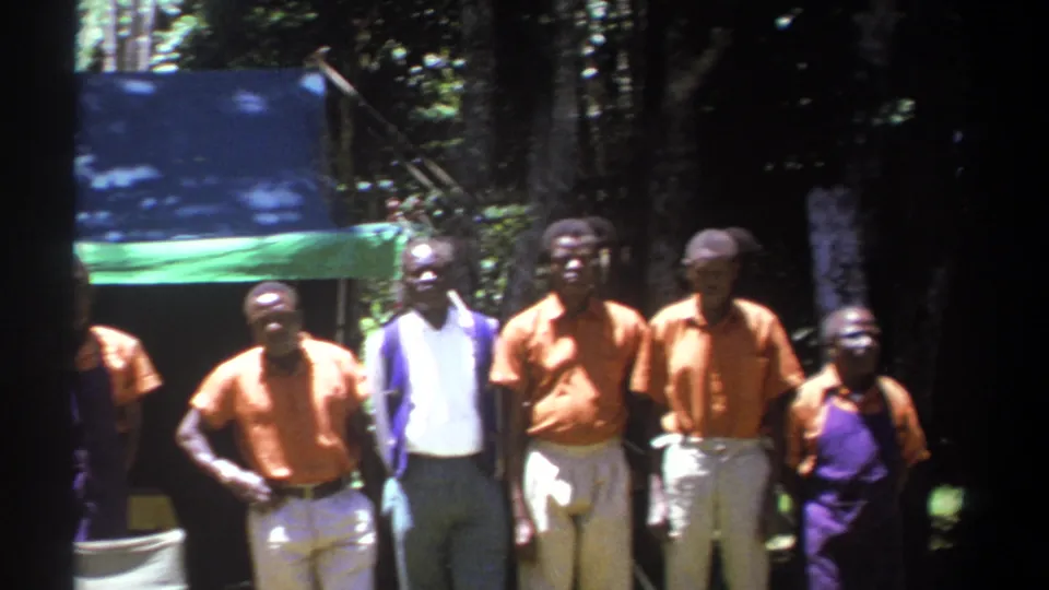 Men in orange shirts, light trousers, and work clothing stand at the edge of a shaded tarp area in Kenya, 1969.