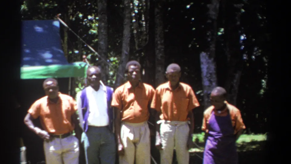 In Kenya 1969 footage, several African men stand in a line beneath a blue and green tarp with trees behind them.