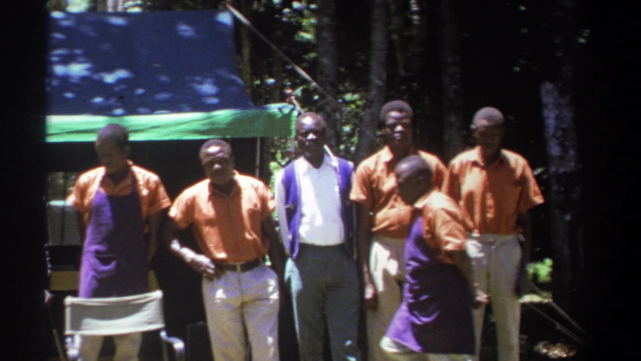 A group of men stands near a tarp, with one man turned sideways in front.