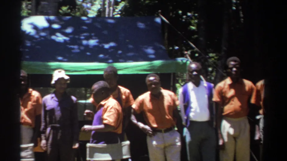 The 1969 Kenya home-movie frame shows men gathered in shade beneath a tarp, with several orange shirts and purple garments visible.