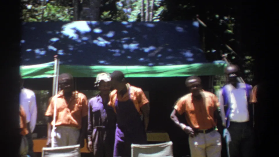 In this Kenya 1969 frame, men in orange shirts and purple aprons stand near a folding chair under a tarp in filtered sunlight.