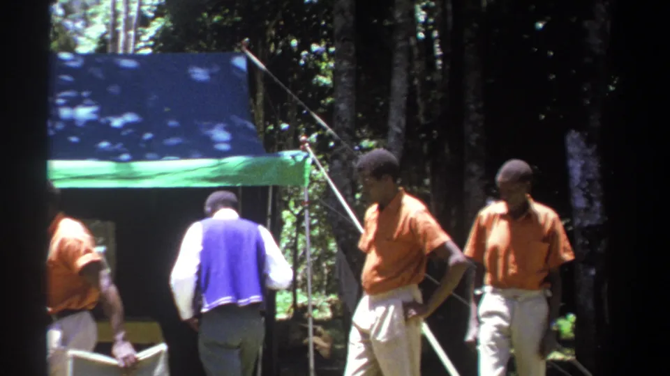 1969 Kenya home-movie frame showing men around a temporary tarp shelter, with chairs and trees visible behind them.