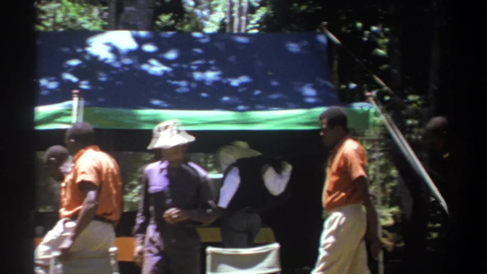 1969 Kenya frame of men in orange shirts and other clothing gathered beside a blue and green tarp shelter in woodland.