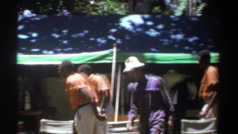 In Kenya 1969 home-movie footage, men in professional-looking clothes pass a green and blue tarp shelter among trees.