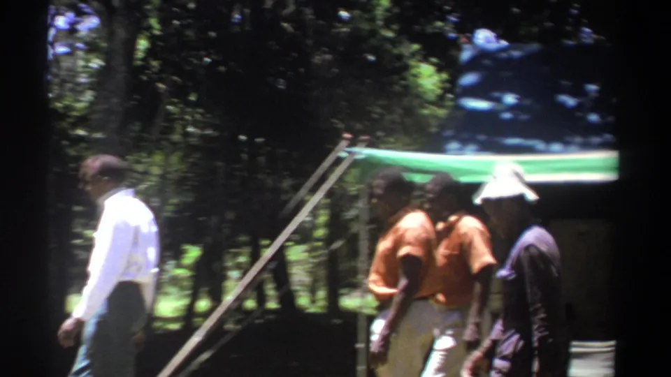 A man in a white shirt passes near a group of men by a tented structure in wooded Kenya, filmed on 8mm in 1969.