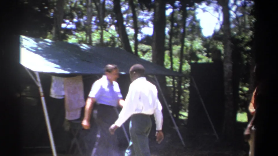 Two adult men move near a canvas shelter in a wooded Kenyan camp scene, with other figures partly visible at the edges.