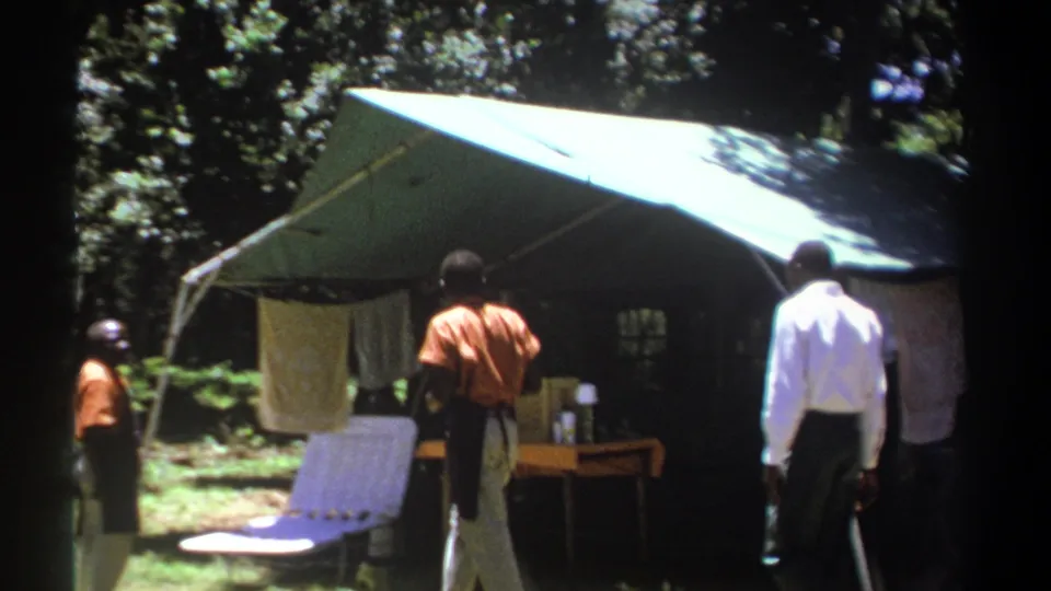 Men in a wooded Kenya campsite stand near a green tarp shelter, with a white chair and small table visible beneath the awning.