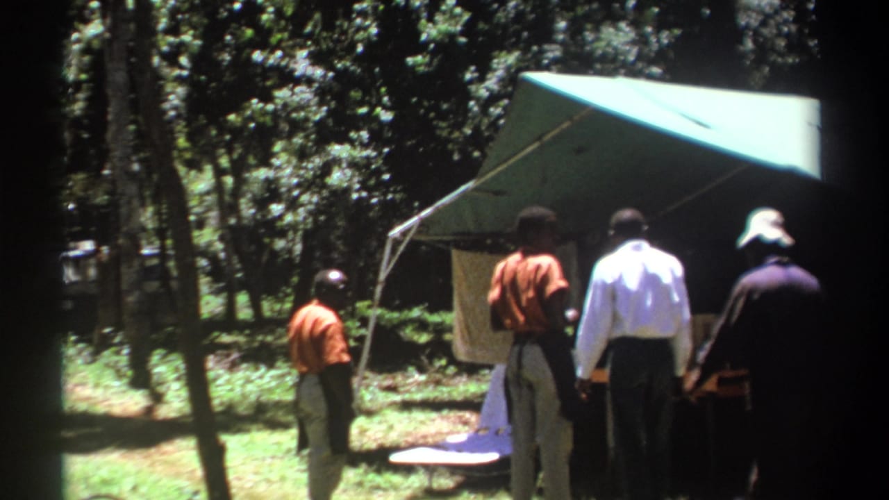 Several men stand beside a tent and table under trees.