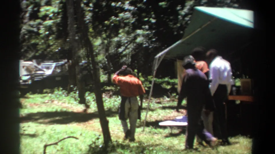 1969 Kenya home-movie frame showing men moving through a forest campsite with a vehicle in the background.