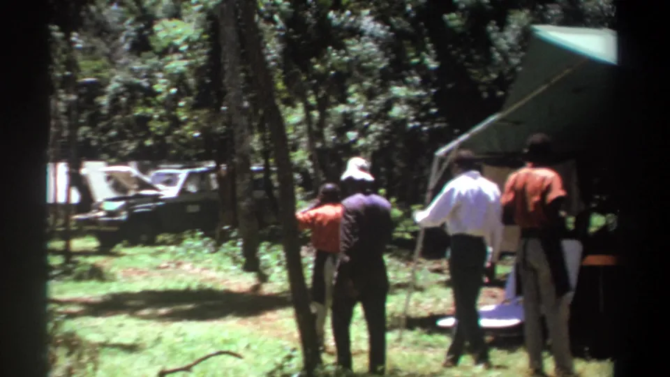 1969 Kenya frame showing men at a wooded campsite, with a tent and a vehicle partly visible behind trees.