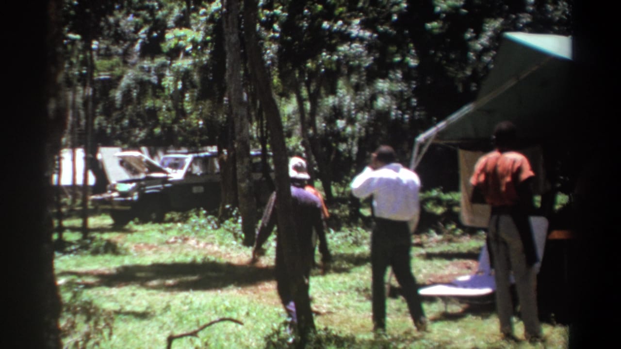 Men walk through a wooded campsite near a green tent and a vehicle with its hood open.