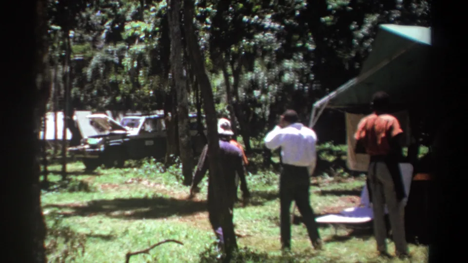 In Kenya in 1969, several men walk away from a green tent through a sunlit wooded campsite with a vehicle in the background.