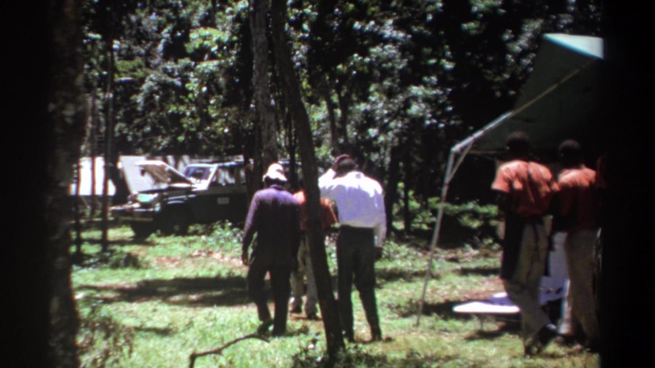 A group of men continues walking past trees toward a campsite vehicle and tent.