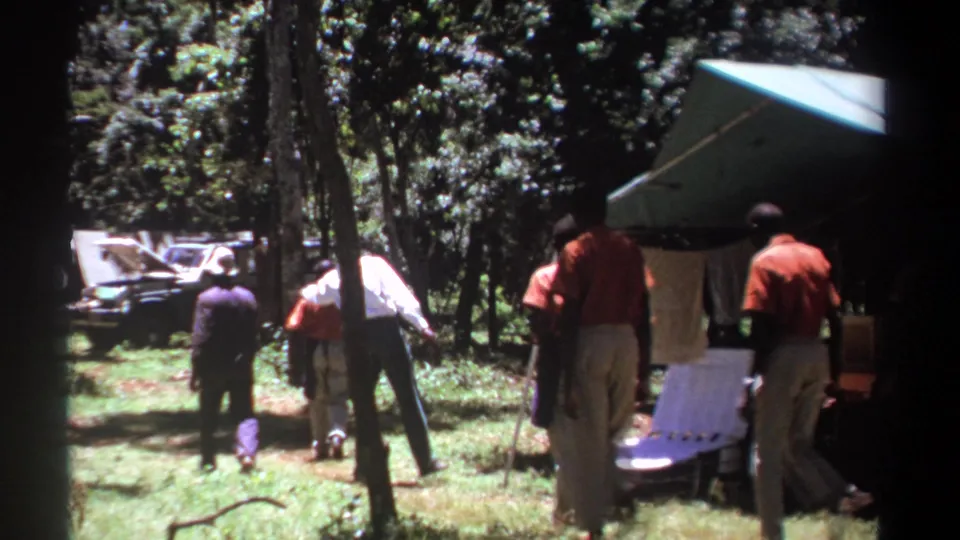The 1969 Kenya home-movie frame shows men walking away from the camera beside camp furniture and a green tent.