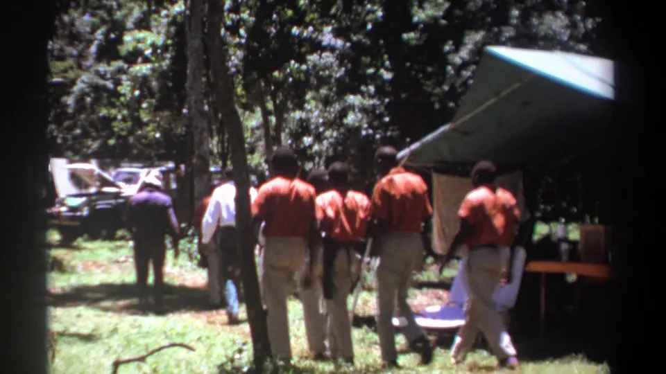 Several men in matching shirts move through the wooded Kenya campsite, with a tent, table, and vehicle visible behind them.