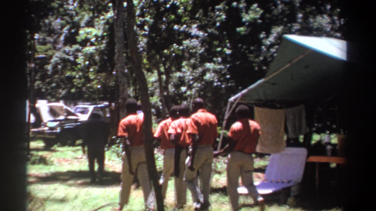 Men in orange shirts walk through a wooded camp beside a canvas shelter.