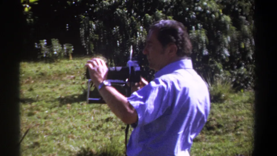 1969 Kenya 8mm frame of a man in a blue shirt using a larger camera in a grassy area bordered by trees.