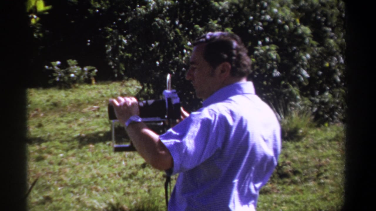 Man in a pale blue shirt holds a vintage film camera outdoors near grass and trees.
