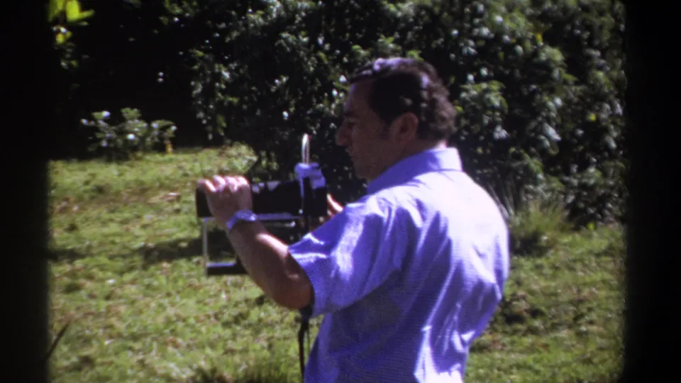 In Kenya-dated 1969 home-movie footage, a man in a pale blue shirt holds a vintage 8mm camera in a grassy wooded area.