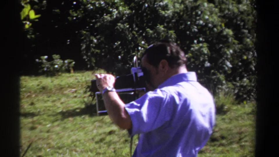 A man in Kenya-dated 1969 footage appears to adjust a vintage 8mm camera while standing beside grass and dark green trees.