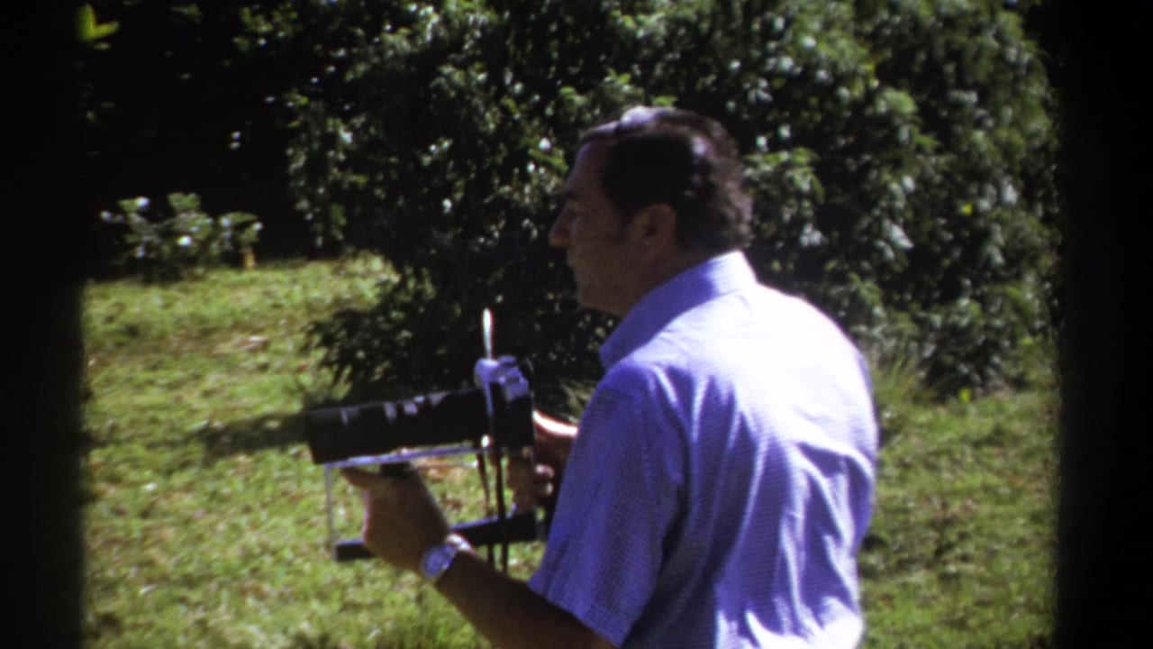 Man in a light blue shirt holds a vintage movie camera outdoors among grass and trees.