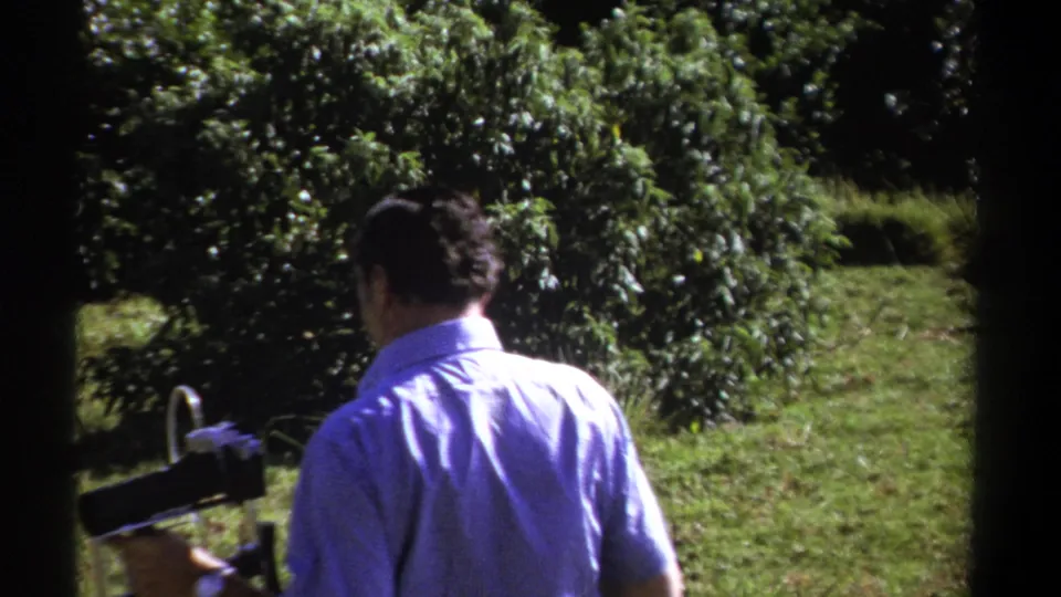 The camera operator is seen from behind in the Kenyan outdoor setting, carrying a vintage cine camera beside sunlit grass and shrubs.