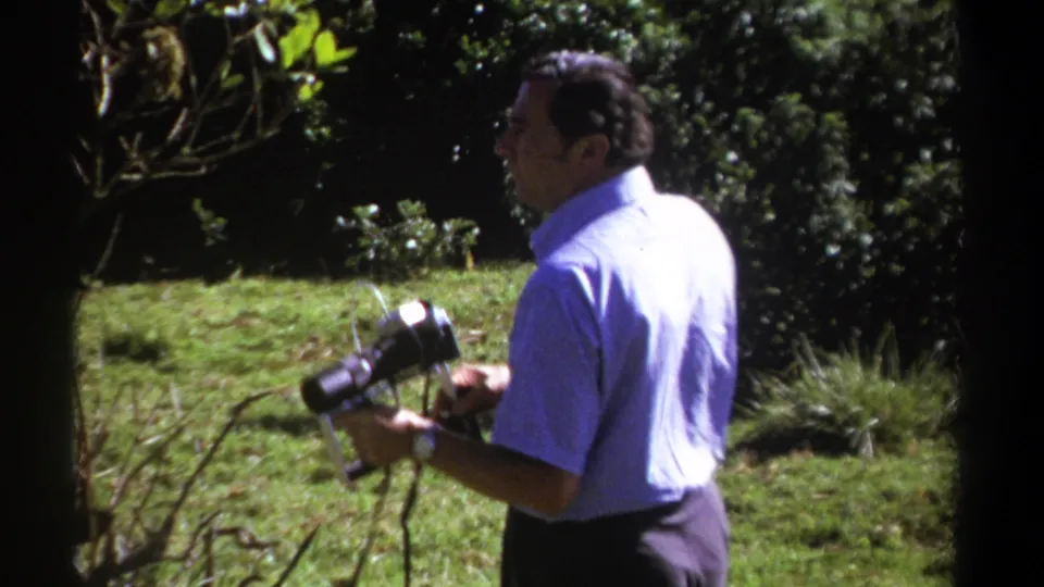 A man in a short-sleeve shirt holds a vintage cine camera near branches, grass, and trees in 1969 Kenya footage.