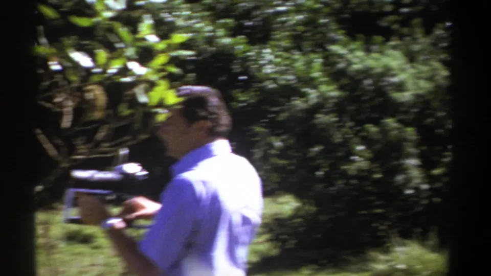 A motion-blurred frame shows the man and vintage camera moving past dense foliage in the Kenyan outdoor scene.