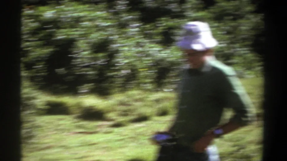 In 1969 Kenya footage, a man in a white bucket hat and green shirt moves through a wooded clearing with heavy motion blur.