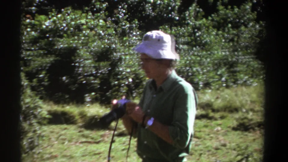 In 1969 Kenya footage, the man stands in profile in a green clearing while holding a compact camera by its strap.