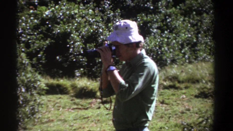 In 1969 Kenya footage, the man holds the camera to his eye, framed by sunlit grass and thick green foliage.