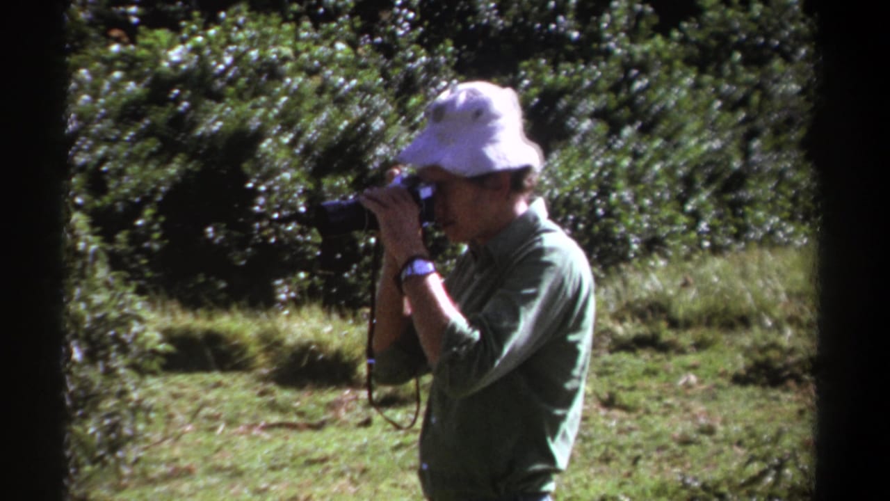 Man in a white bucket hat holds a camera to his face in a grassy clearing.