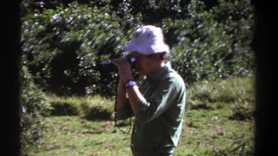 In Kenya 1969 home-movie footage, a man in a white bucket hat and green shirt aims a still camera outdoors.