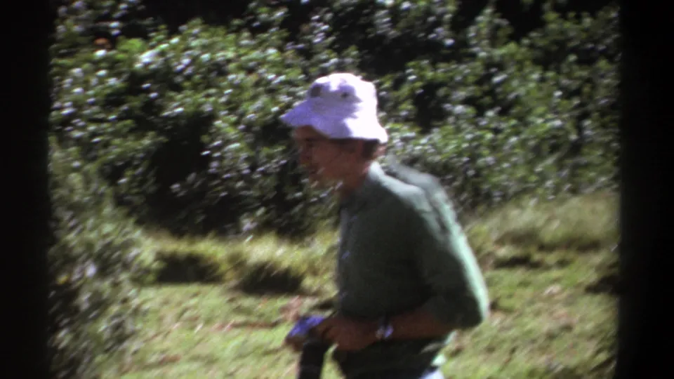 The photographer lowers his camera in the Kenya 1969 home-movie scene, with trees and grass behind him.