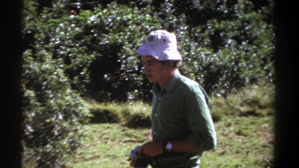 In 1969 Kenya footage, a man in a white bucket hat and green shirt stands in bushland holding a still camera with a long lens.