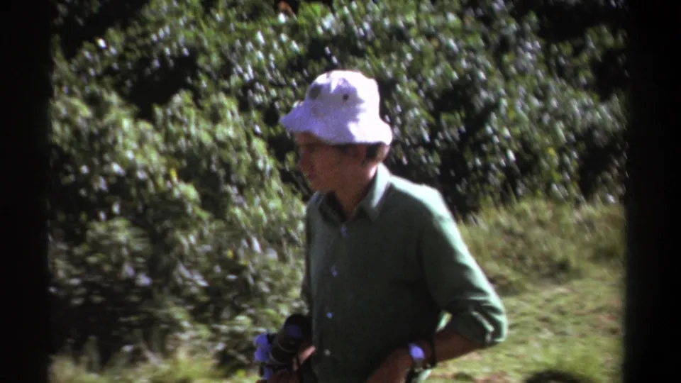 The man appears to check or steady his camera while standing against dense green bushland in the Kenya home-movie footage.