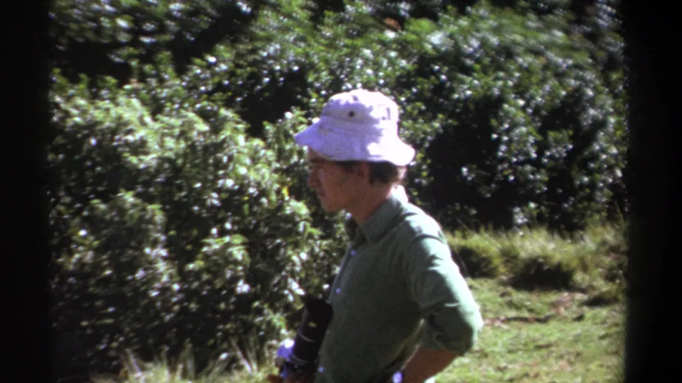 The 1969 Kenya frame shows the man in profile, holding camera equipment while facing into a sunlit area of shrubs and grass.