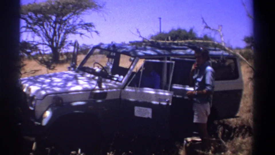 1969 Kenya home-movie frame showing a man in shorts beside a dark off-road vehicle with brush on the roof rack.