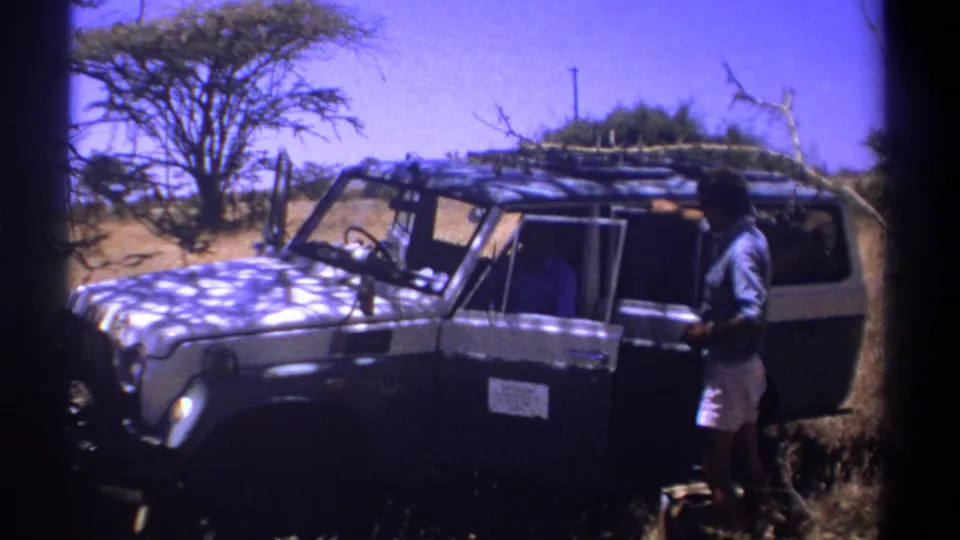 1969 Kenya frame of a man standing by the passenger side of a roof-loaded off-road vehicle in dry open terrain.