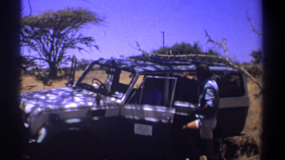 1969 Kenya home-movie still with a man beside a dark off-road vehicle, sparse trees, and dry roadside vegetation.