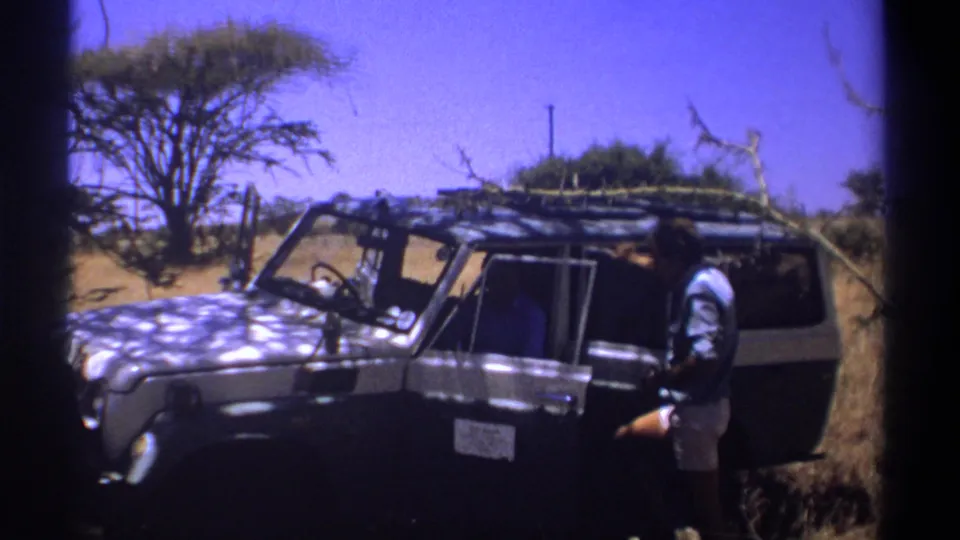 1969 Kenya frame showing a man next to a roof-racked off-road vehicle, with dry grass and scattered trees behind him.