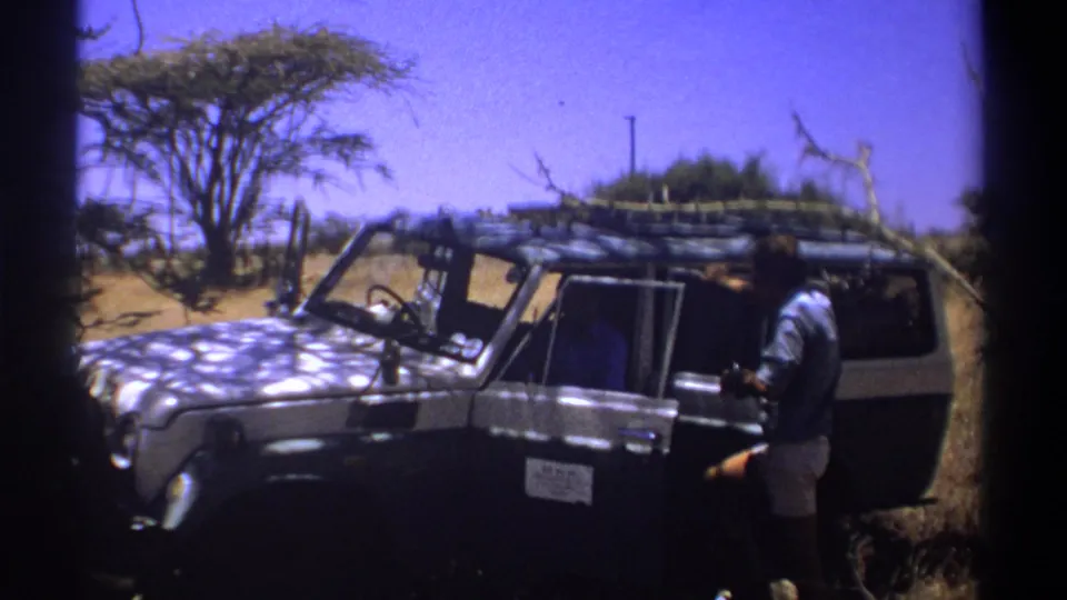 Kenya 1969 frame of a man in light shirt and shorts near a parked safari vehicle, with dry brush and a flat blue sky in the background.