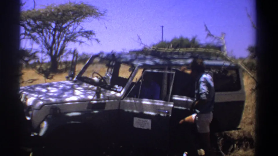 Kenya 1969 home-movie still with a dark safari vehicle, open passenger-side door, roof rack, and a man standing in dry savanna light.