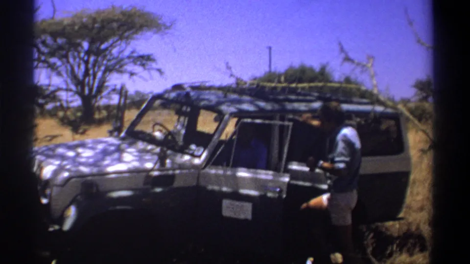 Kenya 1969 frame showing a man at the side of a roof-rack safari vehicle, apparently interacting with someone inside, against dry savanna vegetation.