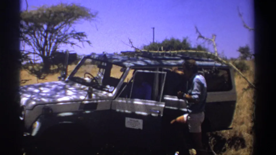 In Kenya in 1969, a man stands beside a dark 4x4 safari vehicle with a roof rack under bright sun.