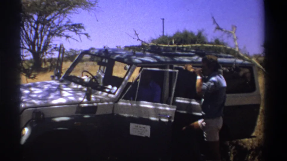 A man in light shorts and a blue shirt stands beside a 1969 Kenya safari vehicle in a dry landscape.